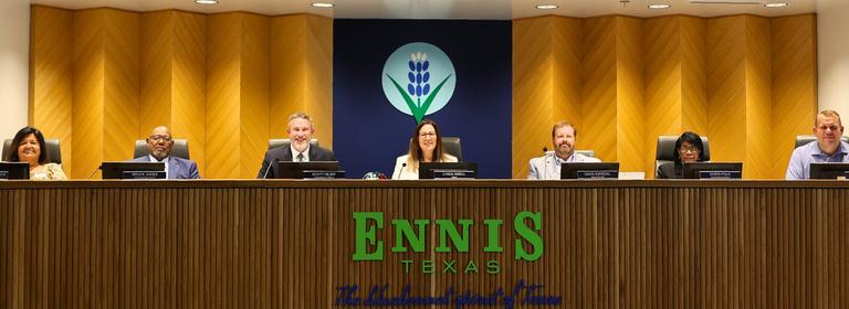 Group of individuals seated behind a long wooden dais in a meeting room, with a decorative emblem featuring a stylized blue flower on the wall behind them and the words ‘Ennis Texas’ displayed on the front of the dais.